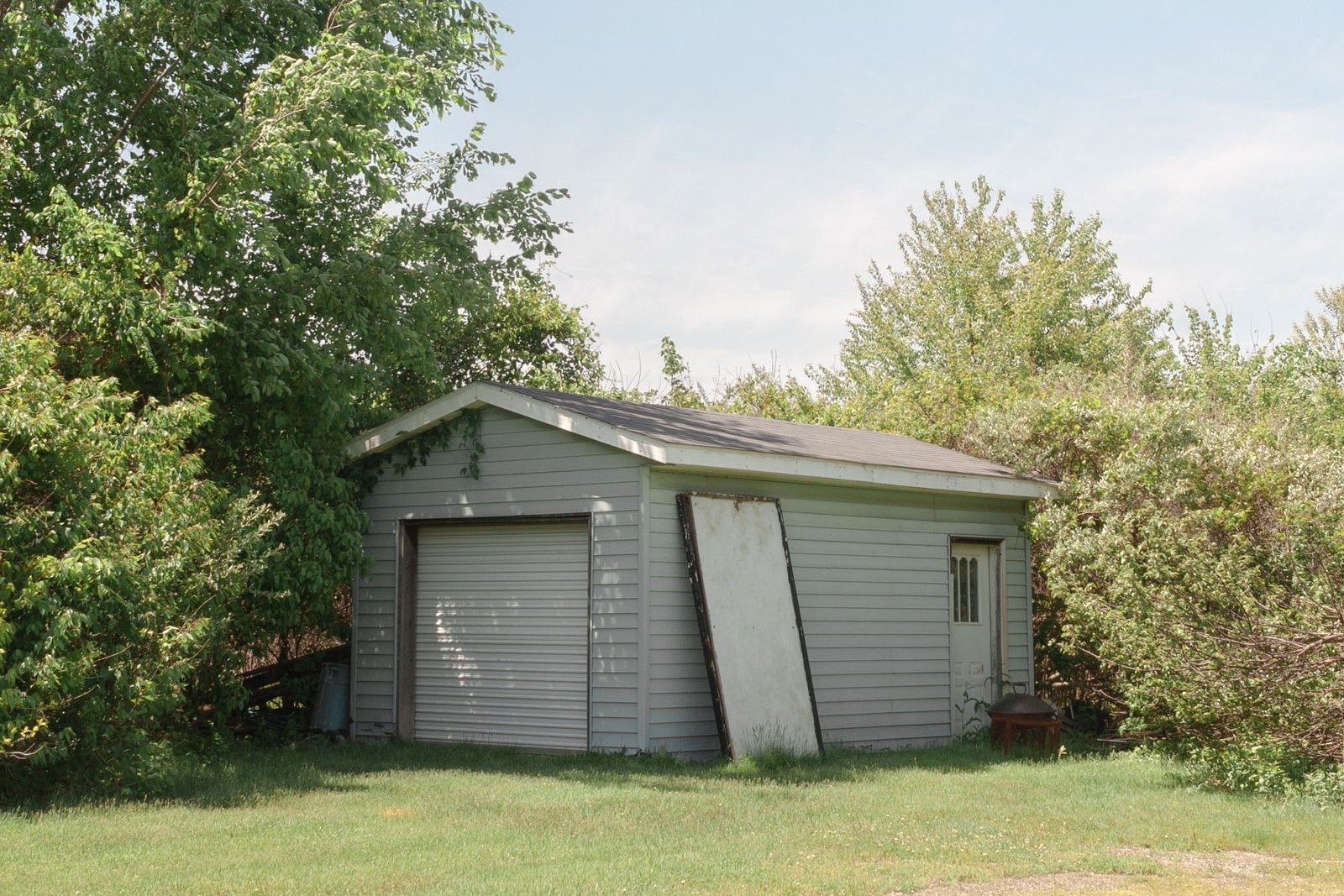 A small detached garage with a roll-up door and pale siding standing at the edge of a mowed yard, surrounded by trees under a hazy summer sky