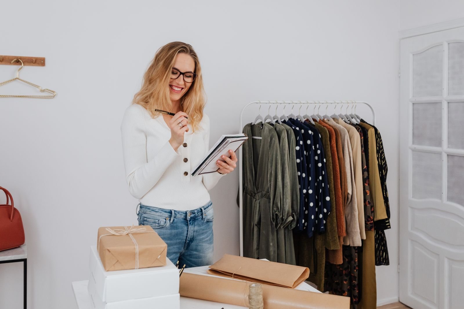 A smiling shop owner in glasses writing in a notebook next to a rack of clothing and a stack of wrapped packages in a small boutique