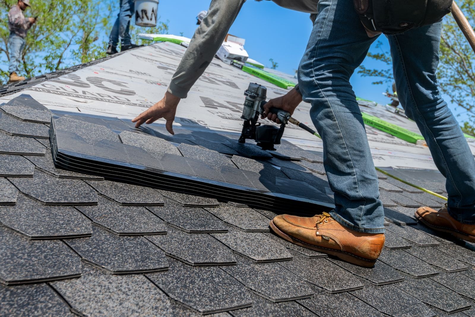 A roofing crew laying asphalt shingles over underlayment on a steep roof, with a coil nailer and a worker's boots in the foreground