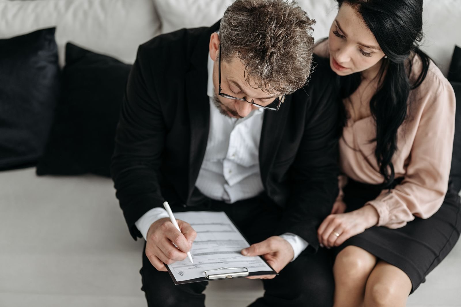 A man and woman seated on a sofa, reading and signing a document on a clipboard together