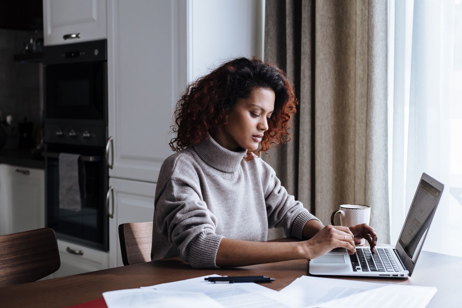 A woman working on a laptop at a kitchen table with papers and a coffee mug beside her